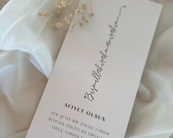 May include: A rectangular card with text in a script font, with the words "Bismillahirahmanirahim" at the top. The card also has the words "AFIYET OLSUN" and other text in a script font. The card is light pink and is decorated with dried flowers.