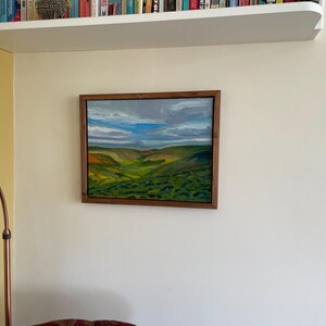 K&ouml;nnte beinhalten: Gerahmtes Landschaftsgem&auml;lde mit einem Tal mit gr&uuml;nen H&uuml;geln, gelben und orangen Akzenten und einem bew&ouml;lkten blauen Himmel. Das Kunstwerk befindet sich in einem dunklen Holzrahmen und h&auml;ngt an einer wei&szlig;en Wand.