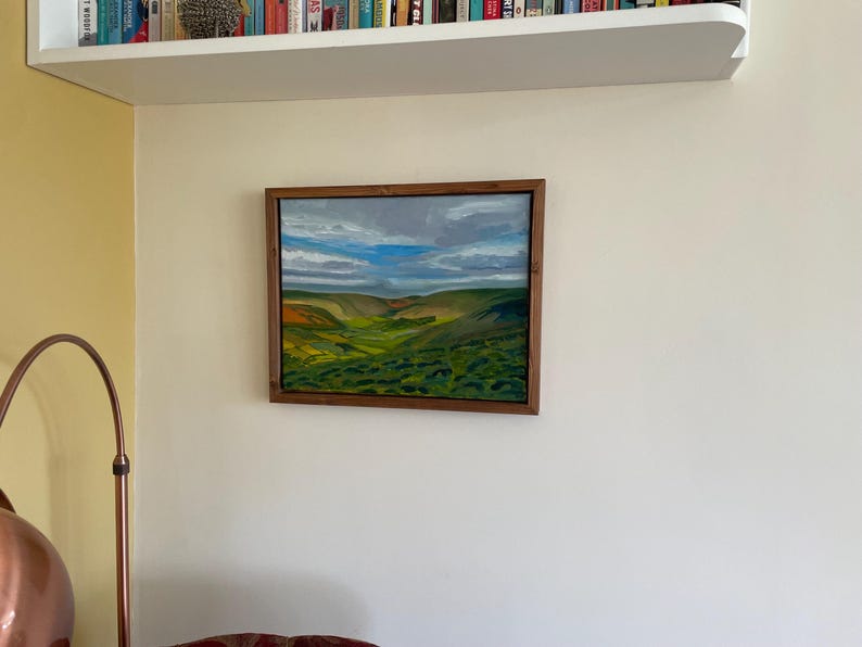 K&ouml;nnte beinhalten: Gerahmtes Landschaftsgem&auml;lde mit einem Tal mit gr&uuml;nen H&uuml;geln, gelben und orangen Akzenten und einem bew&ouml;lkten blauen Himmel. Das Kunstwerk befindet sich in einem dunklen Holzrahmen und h&auml;ngt an einer wei&szlig;en Wand.