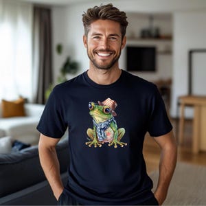 Könnte beinhalten: Marineblaues T-Shirt mit einer detaillierten Illustration eines Frosches, der einen gestreiften Hut und einen blauen Schal mit weißen Sternen trägt. Der Frosch hat große Augen und sitzt aufrecht. Das Shirt ist ein klassischer Rundhalsausschnitt.