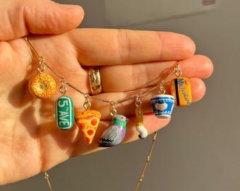 Puede incluir: Un collar dorado con dijes en miniatura de un bagel, un letrero "5 AVE", una porción de pizza, una paloma, un pincel, una taza de café y una MetroCard. Los dijes son coloridos y detallados, con un diseño caprichoso.