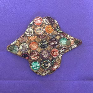 May include: A brown cloth pad with a pattern of vintage bottle caps. The bottle caps have various colors and text, including "Root Beer", "Black Cherry", "Grapefruit", "Lemon Soda", "Lime Soda", and "Punch".