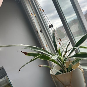 Puede incluir: Una maceta de cerámica blanca con una planta verde colgando de una cadena dorada con cuentas ámbar y transparentes.