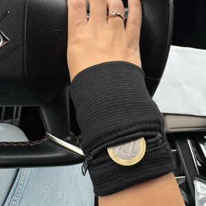 Può includere: Portafoglio da polso nero con tasca con cerniera, contenente una moneta. Il cinturino è realizzato in tessuto strutturato e viene indossato al polso di una persona. La persona tiene un volante nero.