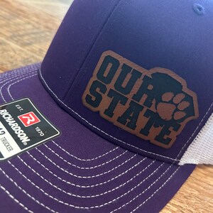 May include: Purple and white trucker hat with the text "OUR STATE" and a paw print design on a brown leather patch. The hat has a curved brim and a mesh back. The tag reads "RICHARDSON 112 TRUCKER".