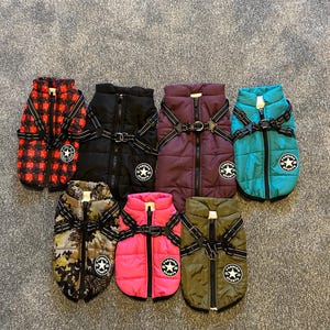 May include: Assortment of eight dog coats in various colors and patterns, including red plaid, black, purple, teal, camouflage, pink, and olive green. Each coat features a black zipper, black straps, and a white star logo with the text "Fashion Sports".