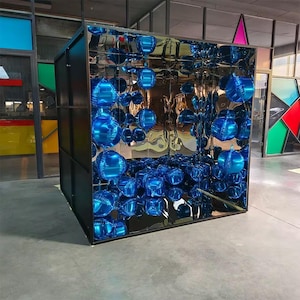 May include: A large, black-framed cube structure featuring mirrored interior walls that create a reflective, infinite effect. The interior is filled with numerous glossy, round, metallic blue balloons, which are multiplied by the mirrors.