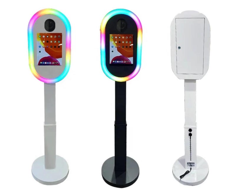 May include: Three photo booth stands, one with a white frame and white stand, one with a black frame and black stand, and one with a rainbow-colored frame and white stand.