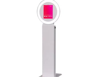 Può includere: Fotomaton bianco con luce ad anello e display touch screen. Lo schermo mostra un gradiente rosa e rosso con icone per scattare foto e video.