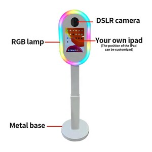 May include: A white photo booth stand with a colorful ring light, a DSLR camera, and a tablet screen. The stand has a metal base.