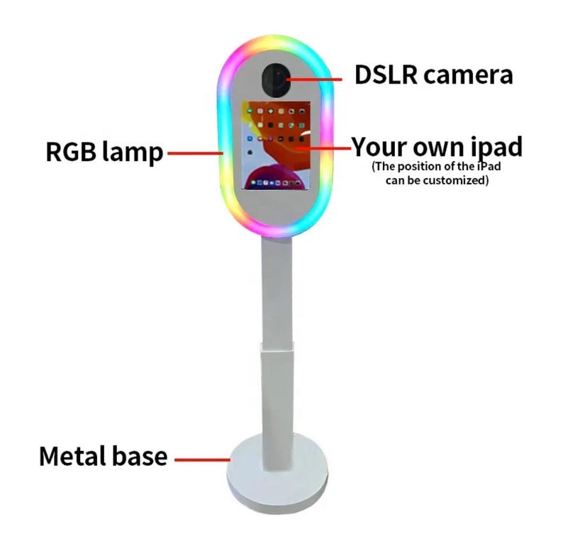 May include: A white photo booth stand with a colorful ring light, a DSLR camera, and a tablet screen. The stand has a metal base.