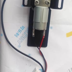 May include: A white electrical box with a black wire attached to it. The box is connected to a black metal bracket with a gray electric motor attached to it. The motor has two red wires and two white wires coming out of it.