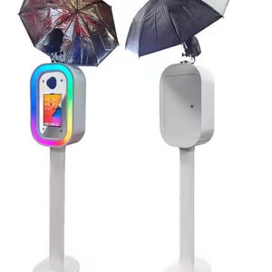 May include: Two white photo booth stands with a black umbrella on top of each. The stand on the left has a colorful light around the photo booth area. The stand on the right has a white door.