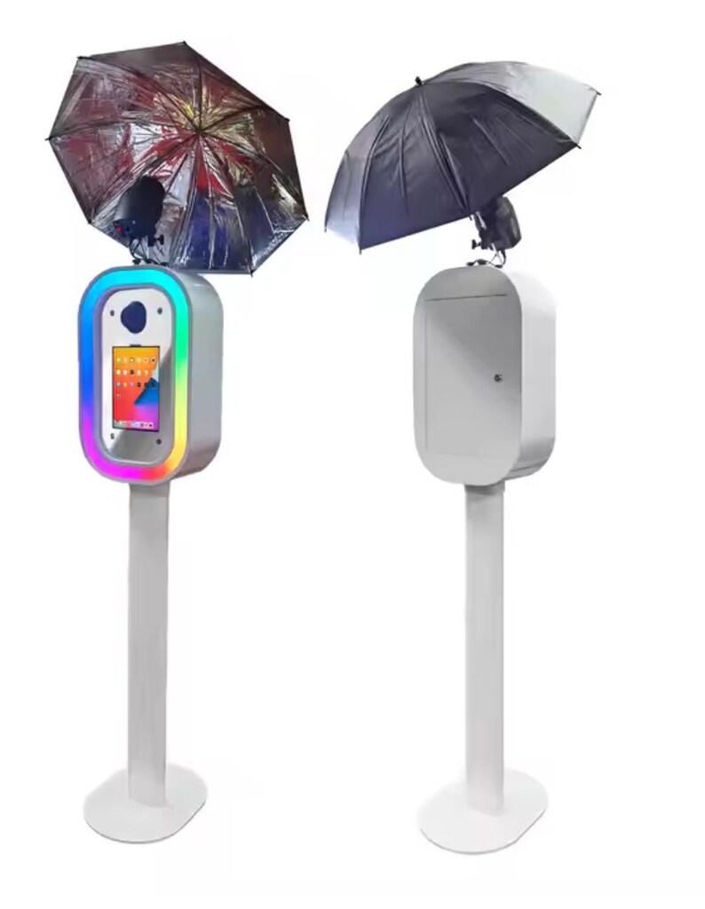 May include: Two white photo booth stands with a black umbrella on top of each. The stand on the left has a colorful light around the photo booth area. The stand on the right has a white door.