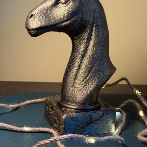 Könnte beinhalten: Eine dunkelgraue Dinosaurierkopfskulptur mit strukturierter Oberfläche. Die Skulptur ist auf einem quadratischen Sockel montiert. Eine Schnur aus kleinen, warmweißen Lichtern ist um den Sockel drapiert.