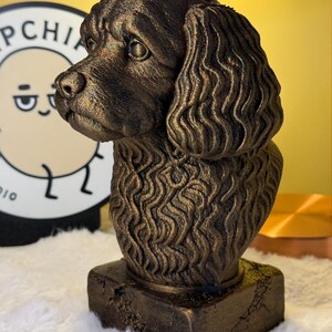 Può includere: Un busto di cane color bronzo con dettagli di pelliccia e tratti del viso. La scultura è montata su una base quadrata con un aspetto strutturato e invecchiato. La testa del cane è girata di lato, mostrando il suo profilo.