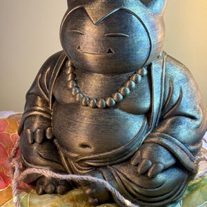 Op de afbeelding: Een bronzen beeld van een stripfiguur in een zittende, meditatieve pose, die lijkt op een Boeddha-figuur. Het beeld heeft een ketting en staat op een kleurrijk patroon. De figuur heeft gesloten ogen en een lichte glimlach.