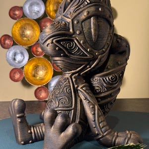 Pode incluir: Estatueta de fantasia de cor bronze de uma criatura semelhante a um sapo em armadura, com detalhes intrincados e um acabamento texturizado. A figura está sentada com as mãos e os pés estendidos. Decoração de parede com esferas metálicas.