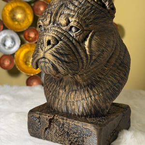 Könnte beinhalten: Eine bronzefarbene Skulptur eines Hundekopfes mit detaillierten Gesichtszügen und einer strukturierten Oberfläche. Die Skulptur ist auf einem rechteckigen Sockel montiert. Der Hintergrund umfasst dekorative Metallkugeln in Gold, Silber und Kupfer.