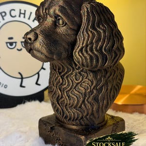 Puede incluir: Busto de perro de color bronce con detalles de pelaje y rasgos faciales. La escultura está montada sobre una base cuadrada y tiene un acabado texturizado. Es un objeto decorativo.