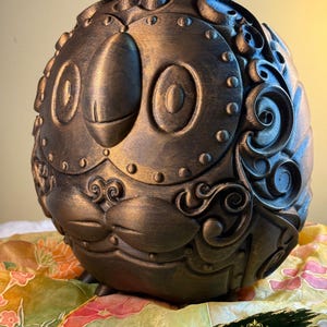 May include: A bronze-colored, spherical sculpture with an owl-like face, featuring large, round eyes and a beak. The surface is detailed with rivets and ornate, swirling patterns. The sculpture is displayed on a floral patterned fabric.
