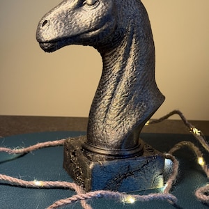 Pode incluir: Uma escultura de cabeça de dinossauro cinza escuro com uma superfície texturizada. A escultura é montada em uma base quadrada. Uma sequência de pequenas luzes brancas quentes é enrolada ao redor da base.