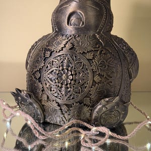 Op de afbeelding: Een bronzen decoratief beeldje met ingewikkelde bloemen- en geometrische patronen. Het beeldje heeft een afgerond lichaam, kleine oren en een serene uitdrukking. Een lichtsnoer is voor het beeldje gedrapeerd.