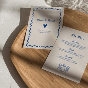 Könnte beinhalten: Zwei weiße Karten mit blauem Text und wellenförmigen Rändern liegen auf einem Holztablett. Auf einer Karte steht "Olivia & Daniel" mit einem Herz und Datum. Die andere ist eine Speisekarte mit "The Menu" oben, mit Vorspeise, Hauptgericht und Dessert.