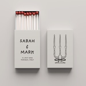 Peut inclure: Une boîte d'allumettes gris clair avec les noms "SARAH & MARK" imprimés en noir, ainsi que la date "12 JUILLET 2026" et le lieu "TOSCANE, ITALIE". La boîte d'allumettes est ouverte, révélant des allumettes à bout rouge. Une deuxième boîte d'allumettes présente un dessin au trait noir d'un chandelier.