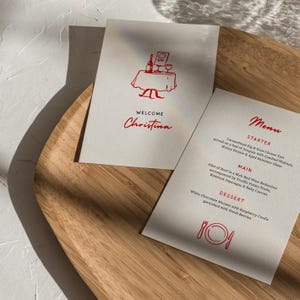 Könnte beinhalten: Zwei weiße Karten mit rotem Text und Illustrationen liegen auf einem Holztablett. Auf einer Karte steht "WELCOME Christina" mit einer Tisch- und Weinflaschenillustration. Die andere ist eine Speisekarte mit Vorspeisen, Hauptgerichten und Desserts.