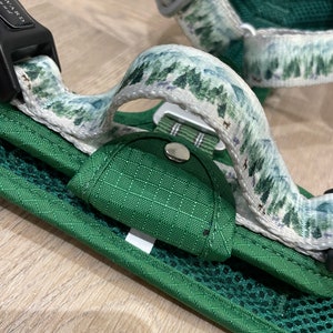May include: Green dog harness with a white strap featuring a forest print. The harness has a black buckle with the word "REWARD" printed on it.