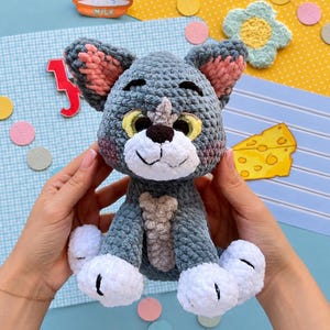 May include: A gray crocheted cat plush toy with white paws, a white muzzle, and large yellow eyes. The cat features pink inner ears, a dark brown nose, and a light gray chest patch. The plush is crafted from soft, textured yarn with a rounded head.