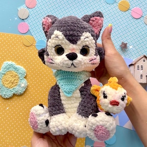 Op de afbeelding: Een donkergrijs en wit gehaakt kattenknuffeltje met grote bruine ogen, roze binnenoren en een lichtblauwe driehoekige halsdoek. Een kleiner oranje en wit gehaakt wezentje is bevestigd aan de zijkant van het lichaam van de kat.