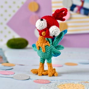 Peut inclure: Figurine d'oiseau crochetée à la main, avec une tête rouge, des yeux blancs et un corps jaune et turquoise. L'oiseau a des pattes orange et des accents bleus sur les ailes. Le fond comprend des découpes de papier colorées.