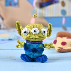 Peut inclure: Un jouet extraterrestre au crochet fait à la main avec trois yeux blancs, une tête vert olive, de grandes oreilles et une petite antenne. L'extraterrestre porte une tenue bleue et violette. Une part de pizza au crochet est en arrière-plan.