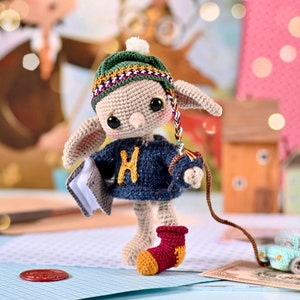 Pode incluir: Uma boneca elfo de crochê vestindo um chapéu verde e amarelo, um suéter azul com um 'H' amarelo na frente e meias vermelhas e amarelas. O elfo está segurando um livro e uma coleira presa a um carro de brinquedo.