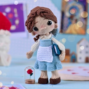 Op de afbeelding: Een gehaakte pop van Belle uit Belle en het Beest, die een blauwe jurk met een wit schort draagt en een boek vasthoudt. De pop heeft bruin haar en bruine ogen. Een glazen stolp met een rode roos erin staat voor de pop.