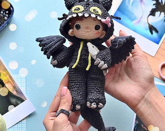 May include: A crocheted black dragon plush toy with yellow accents, wearing a black hooded dragon costume. The toy is being held by a person's hands.
