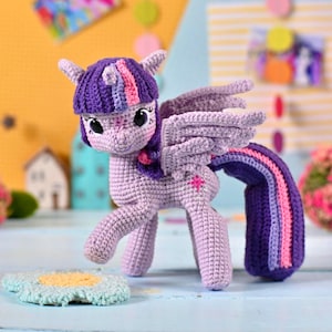 May include: A crocheted lavender pony figurine with large dark eyes, a purple mane featuring pink and blue stripes, and matching wings. The pony has a long, flowing purple tail with pink and blue stripes and a small pink star emblem on its flank.