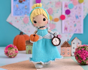 May include: A crocheted Cinderella doll wearing a blue dress and holding a pumpkin and a clock. The doll is standing on a table with a light blue background.