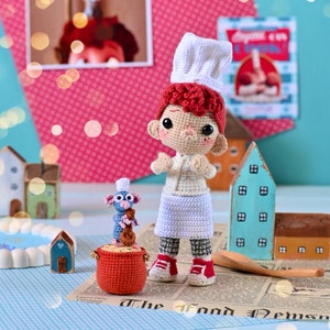 Könnte beinhalten: Eine gehäkelte Chefkochpuppe mit einer weißen Kochmütze und einer weißen und blauen Kochuniform, die neben einer gehäkelten Mauspuppe steht, die ein braunes gehäkeltes Brot hält. Die Puppen stehen auf einer Zeitung mit dem Aufdruck "The Good News".