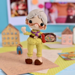 May include: A crocheted doll wearing glasses and a green uniform holds a book that says "My Adventure Book". The doll has gray hair and is standing on a table with papers and a wooden house in the background.