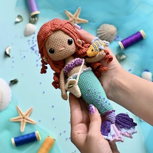 Puede incluir: Una muñeca de sirena de crochet con cabello rojo, un top morado y una cola turquesa. La muñeca sostiene un pequeño pez amarillo de crochet.