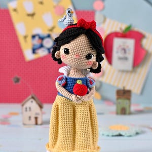 Puede incluir: Muñeca de ganchillo hecha a mano de Blancanieves, con un vestido amarillo, una parte superior azul y cabello negro con un lazo rojo. La muñeca sostiene una manzana roja y tiene un pequeño pájaro azul en la cabeza. La muñeca mide aproximadamente 15 cm de altura.