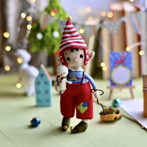 Könnte beinhalten: Eine gehäkelte Elfenpuppe mit einer rot-weiß gestreiften Mütze, einem blauen Hemd und einer roten Latzhose. Der Elf hält einen kleinen gehäkelten Schneemann. Der Elf steht auf einer Holzoberfläche mit einem kleinen Holzhaus und einem Weihnachtsbaum im Hintergrund.