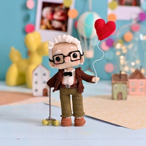 May include: A crocheted amigurumi doll of an older person with white hair, wearing a brown jacket, a white shirt with a bow tie, and brown pants. The doll is holding a walking stick and a red heart balloon.
