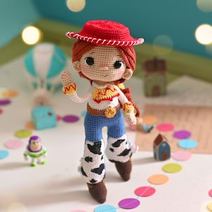 May include: Crocheted toy doll with red hair, a red cowboy hat, a white shirt with a yellow star, blue jeans, and cow print boots. The doll is standing on a white surface with colorful confetti.