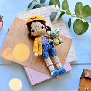 May include: A crocheted doll with yellow hair, wearing a yellow jacket, blue jeans, and a yellow hat with a chick design. The doll is holding a green crocheted frog. The doll is sitting on a stack of papers.
