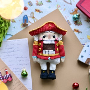 Puede incluir: Un muñeco de cascanueces de ganchillo que lleva un uniforme rojo y dorado con un sombrero blanco. El muñeco tiene la cara pintada de marrón y una gran sonrisa con dientes blancos.