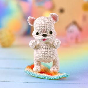 Peut inclure: Une figurine d'ours en crochet blanc cassé, debout sur une planche de surf colorée. L'ours a de petits yeux noirs, un nez et une bouche roses, et est posé comme s'il surfait. La planche de surf est un mélange d'orange, de jaune et de bleu.